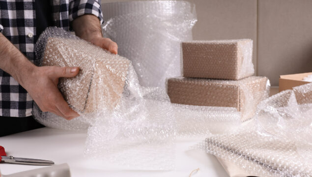 Man covering box with bubble wrap at table in warehouse, closeup