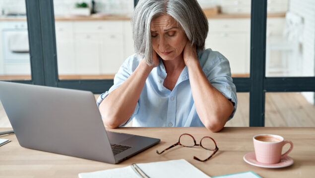 Tired stressed old mature business woman suffering from neckpain working from home office sitting at table. Overworked senior middle aged lady massaging neck feeling hurt pain from incorrect posture.